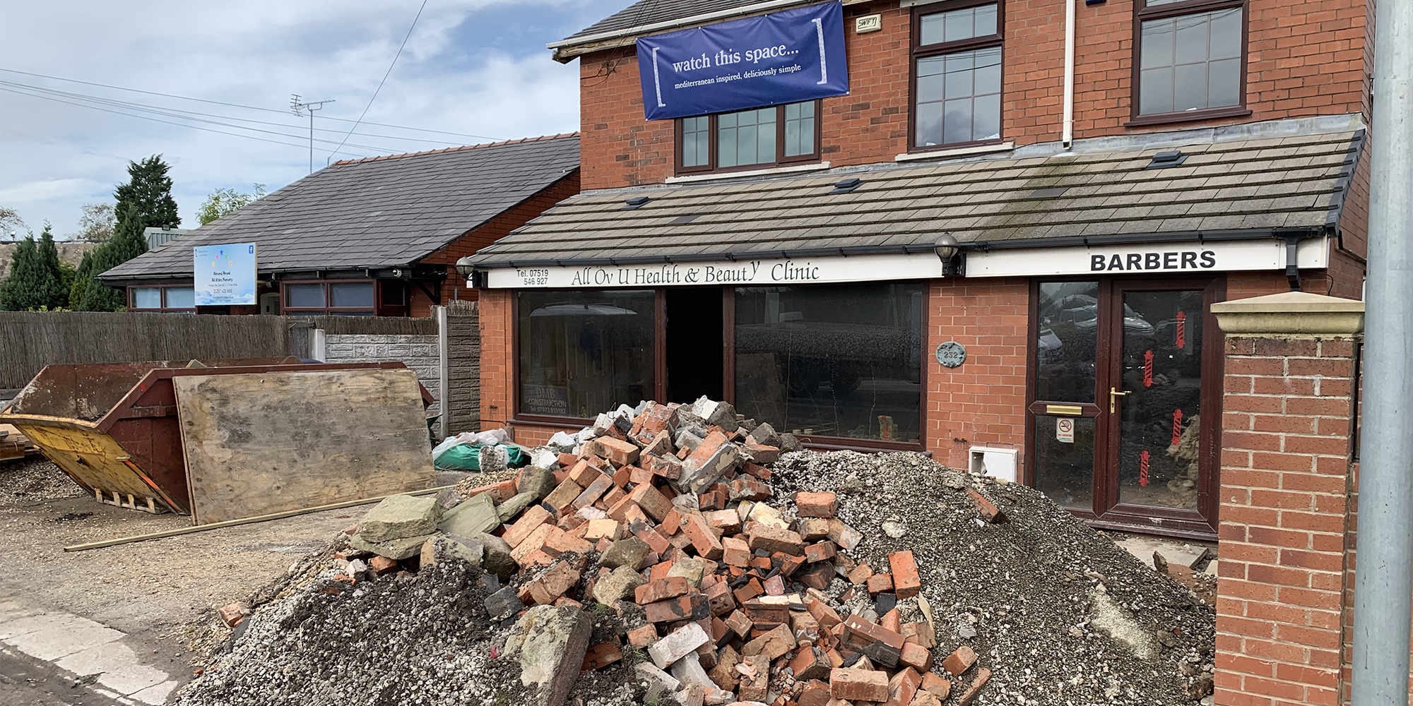 Photo of a neglected building clad only by a 'watch this space' banner with a mound of bricks and rubble in front.